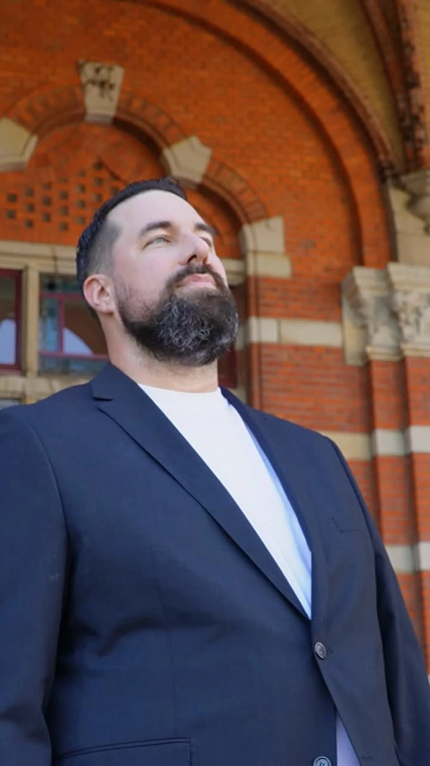 Man met baard en donker colbert staat voor een rood bakstenen gebouw met een grote boogvormige ingang.